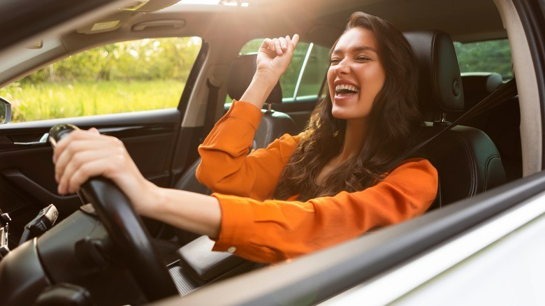 13-a-smiling-woman-in-an-orange-shirt-driving-a-car-looking-joyful-with-one-hand-on-the-steering-wheel-and-the-other-raised-in-excitement
