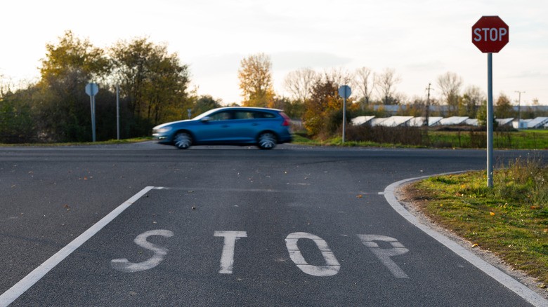8-view-of-a-road-with-a-large-white-stop-painted-on-the-pavement-a-stop-sign-on-the-right-and-a-blue-car-passing-through-the-intersection