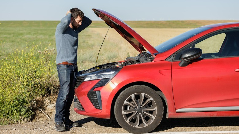 11-a-man-standing-on-the-side-of-the-road-looking-frustrated-holding-his-head-while-examining-the-engine-of-a-red-car-with-the-hood-open